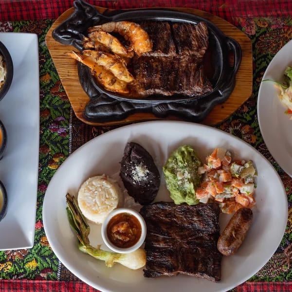 Overhead spread of grilled steak, shrimp, rice, plantains, salads, coleslaw, sauces, and vegetables on colorful woven tablecloth.