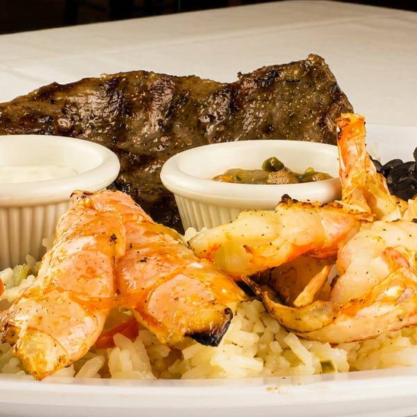 Grilled steak and shrimp with rice, black beans, salad, and two dipping sauces on a white plate.